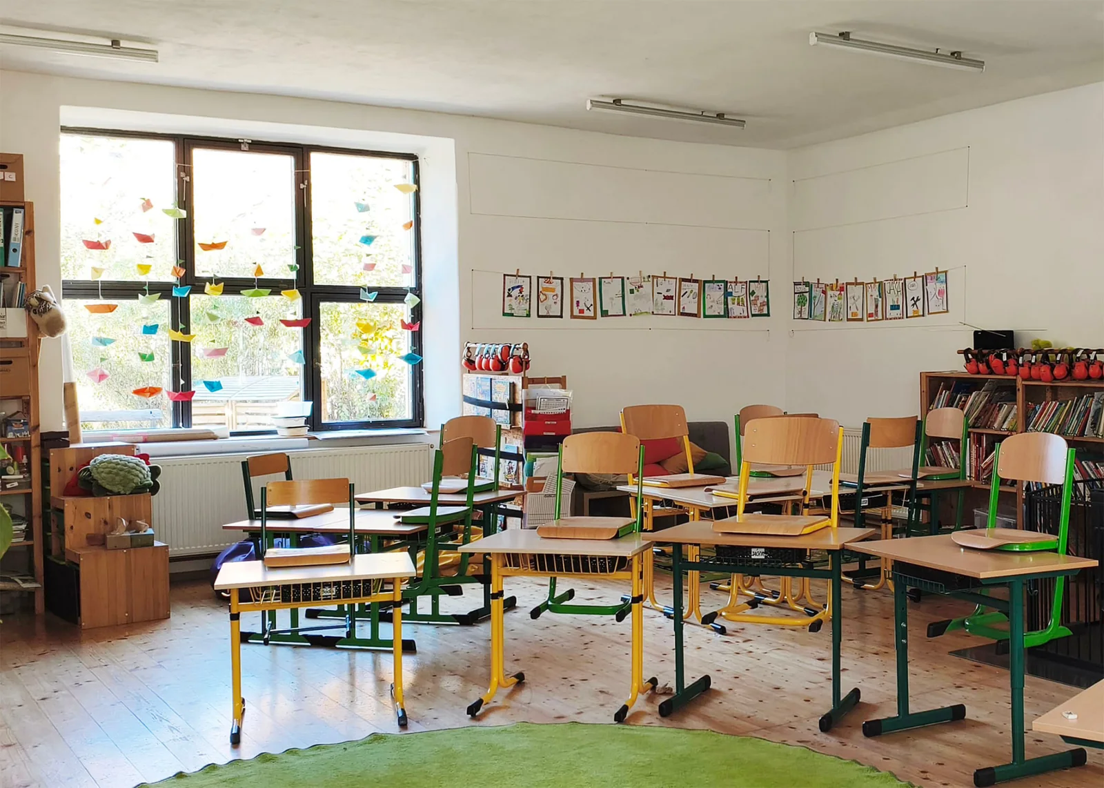 Třída prvního stupně s dřevěnými lavicemi a barevnými papírovými loděčkami zavěšenými ve okně
