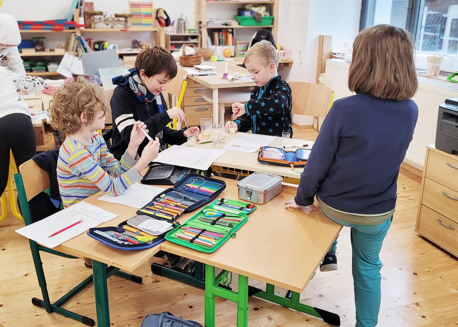 Děti u lavic míchají tekutiny v plastových kelímcích, na deskách mají pencil boxy a pracovní listy